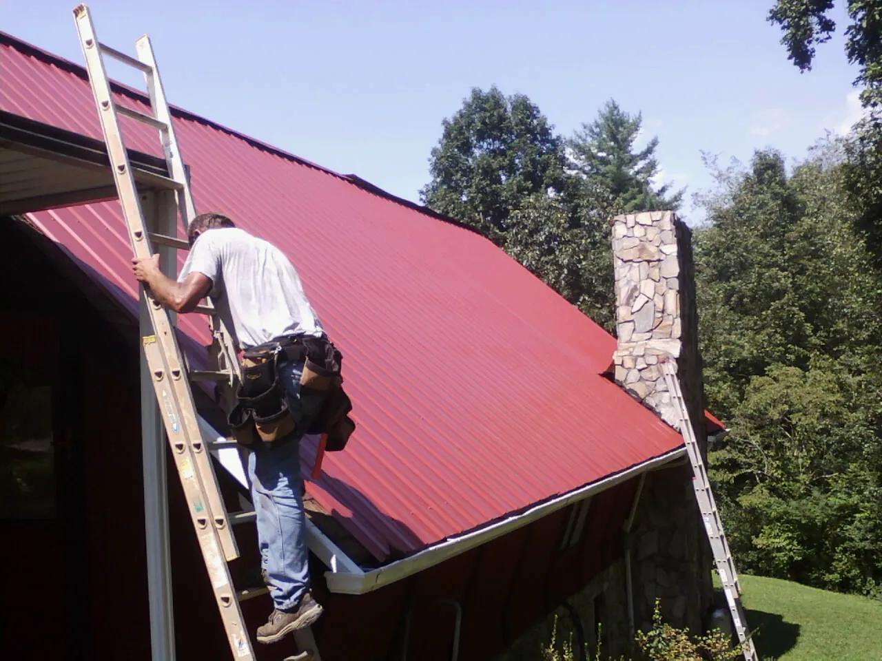 Roofer on a ladder beside a red metal roof and stone chimney — Soffit & Fascia Repair by Roofing Brothers in Red Banks